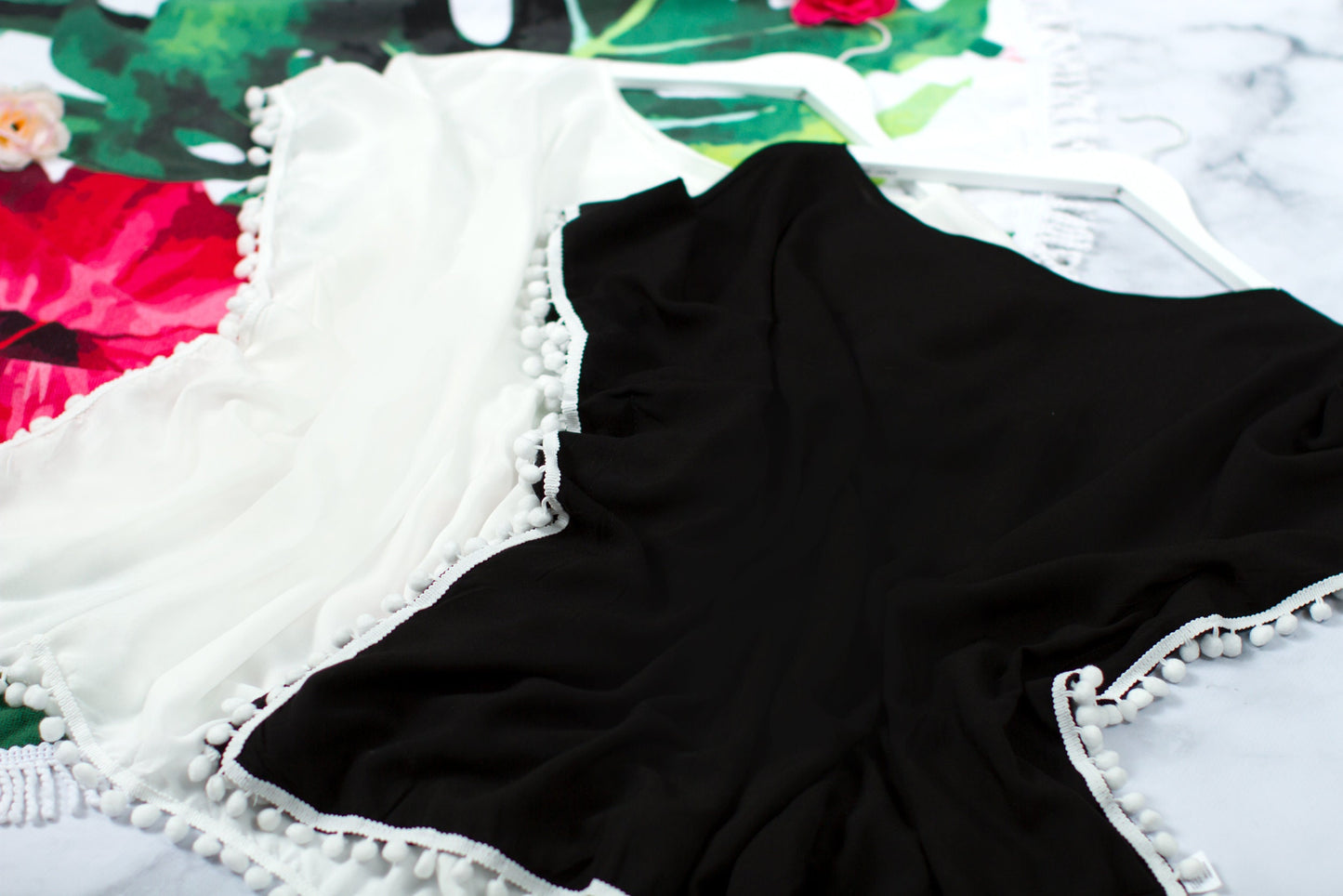 White and black cover-ups folded on marble, showing the pom-pom edging along the hem and sleeves