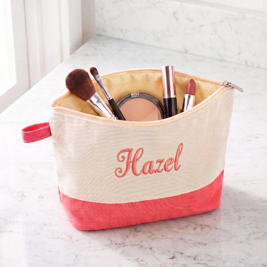 Open cream canvas cosmetic bag with coral-pink base, embroidered Hazel and filled with makeup essentials.
