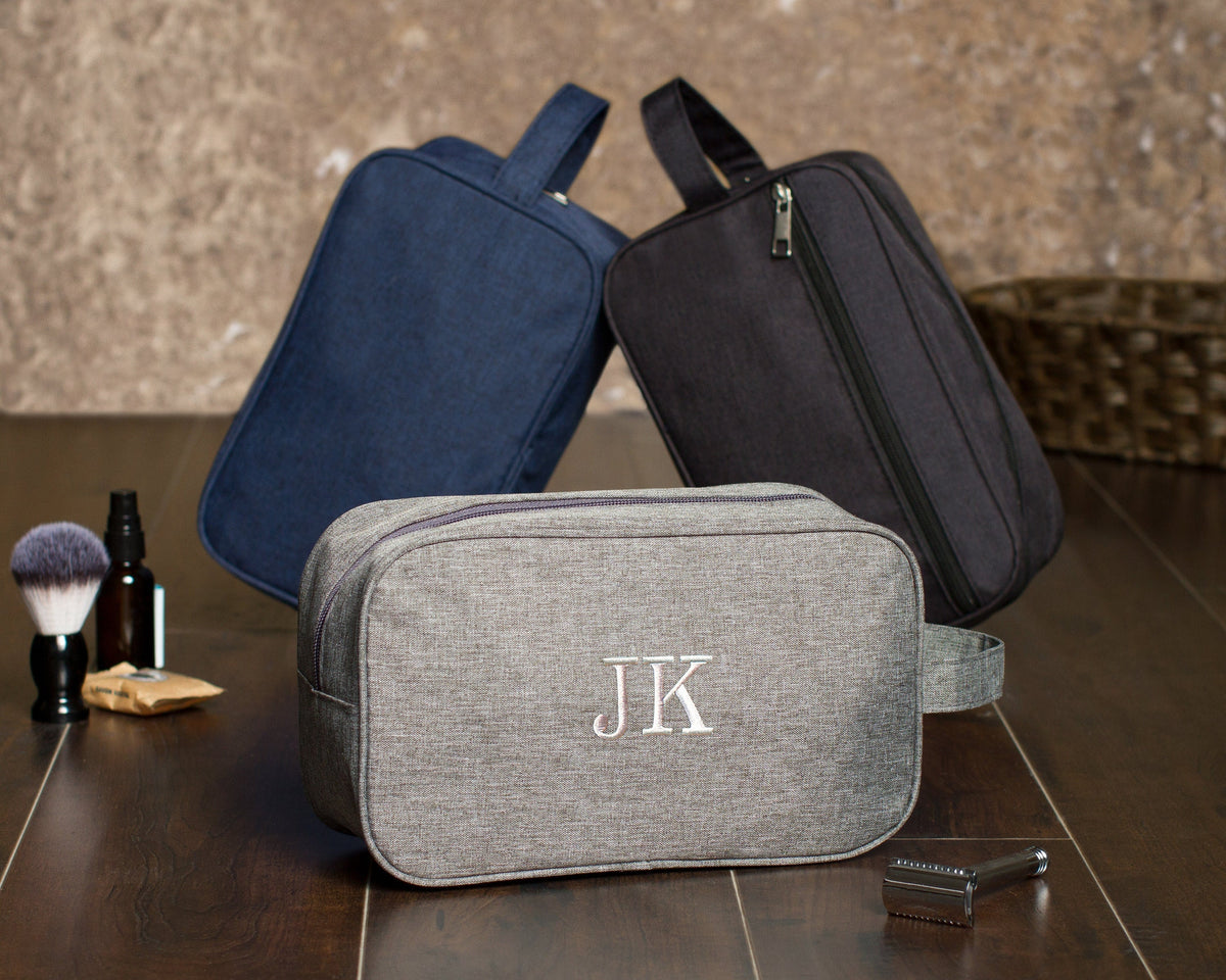 Three heathered canvas dopp kits in navy, charcoal and gray on a wood counter, the gray one embroidered "JK" beside a shaving brush and razor