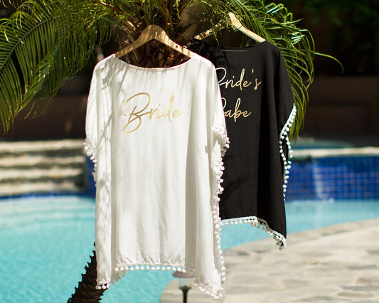 White and black pom-pom beach cover-ups on wooden hangers by a pool, printed "Bride" and "Bride's Babe" in gold