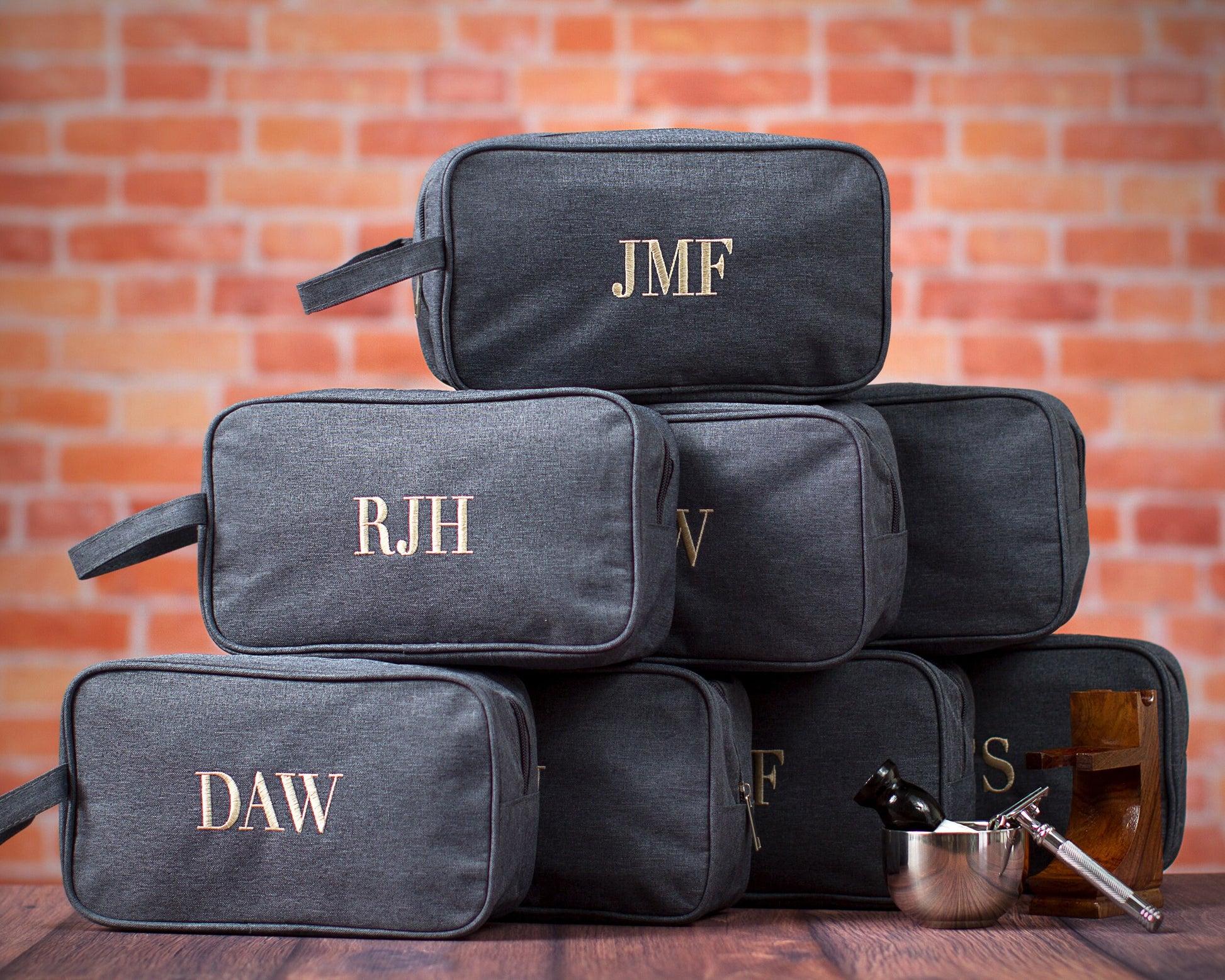 A stack of charcoal dopp kits embroidered with different sets of initials for a groomsmen party, beside a shaving bowl and brush