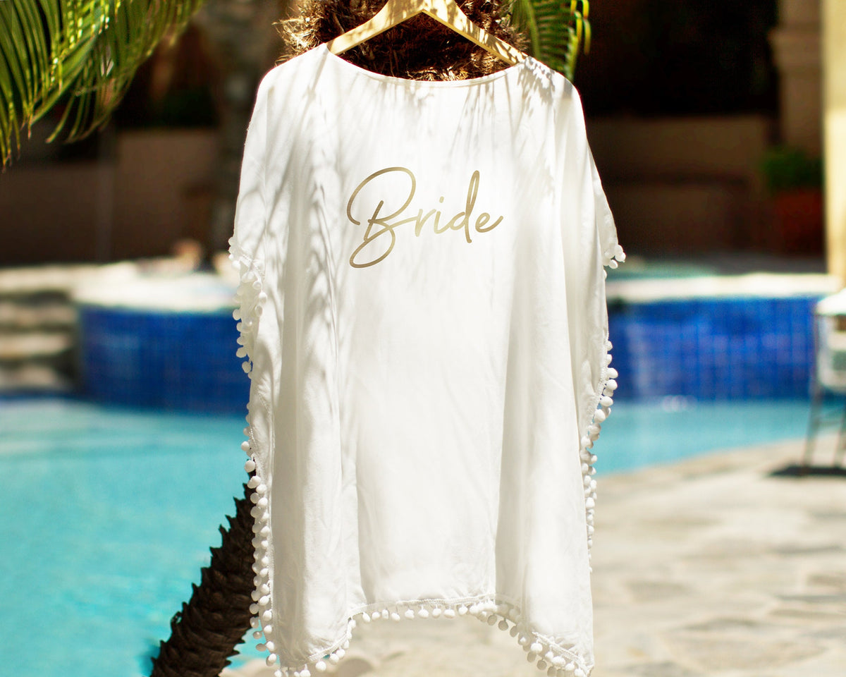 White pom-pom cover-up on a hanger in front of a swimming pool, "Bride" printed across the back in gold