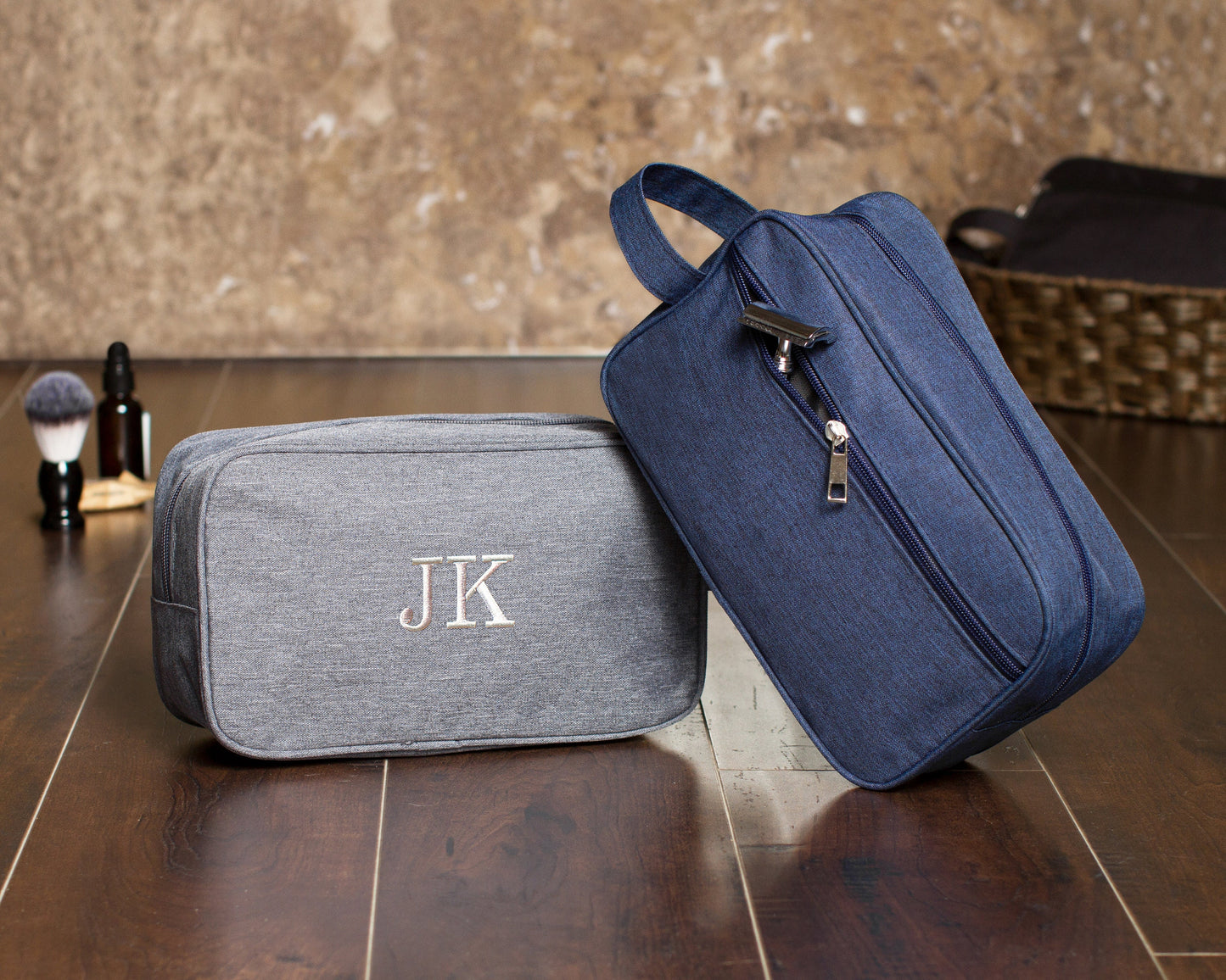 Gray dopp kit embroidered "JK" laid flat beside a navy kit standing on end, showing the loop handle and full-length zipper