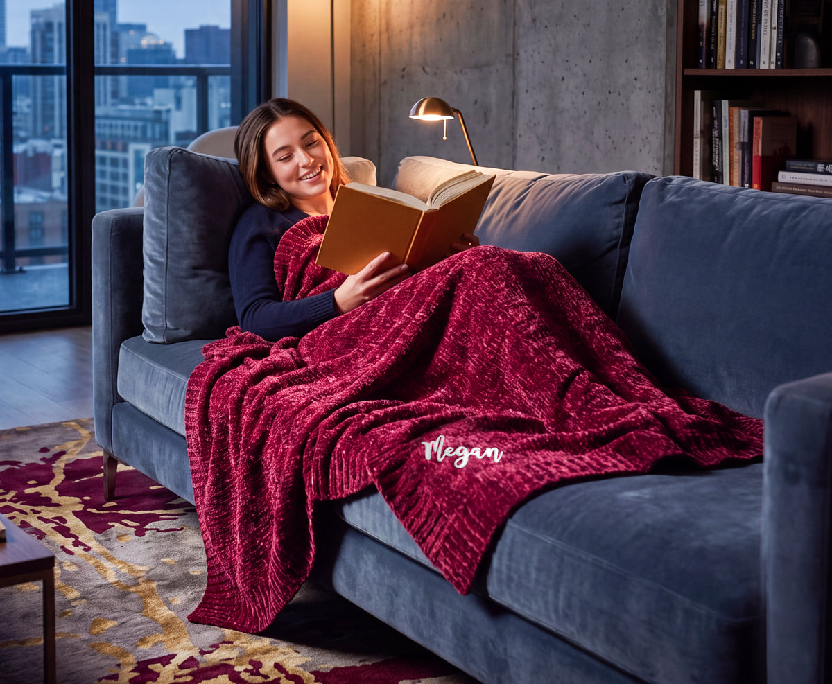 Reader curled up on a sofa under a Nebula burgundy Stargazer throw with Megan embroidered near the edge.