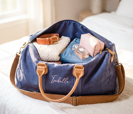 A navy blue canvas duffel bag with tan leather handles and strap sits open on a bed, filled with folded clothing, a pink pouch, a perfume bottle, and a brown belt. The bag has the name 'Isabella' embroidered in white script on the front.
