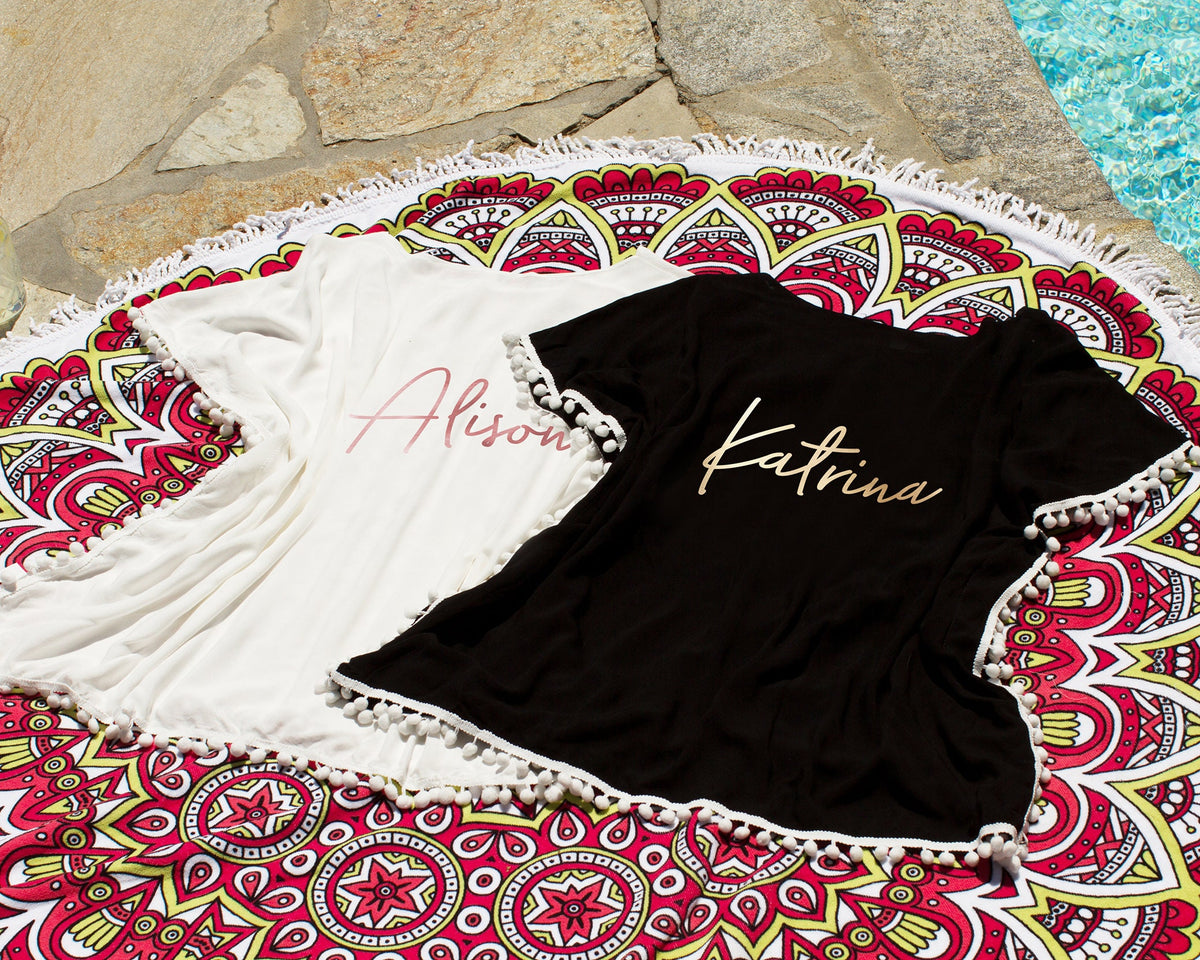 White and black cover-ups laid on a patterned round beach towel, personalized "Alison" in rose gold and "Katrina" in gold