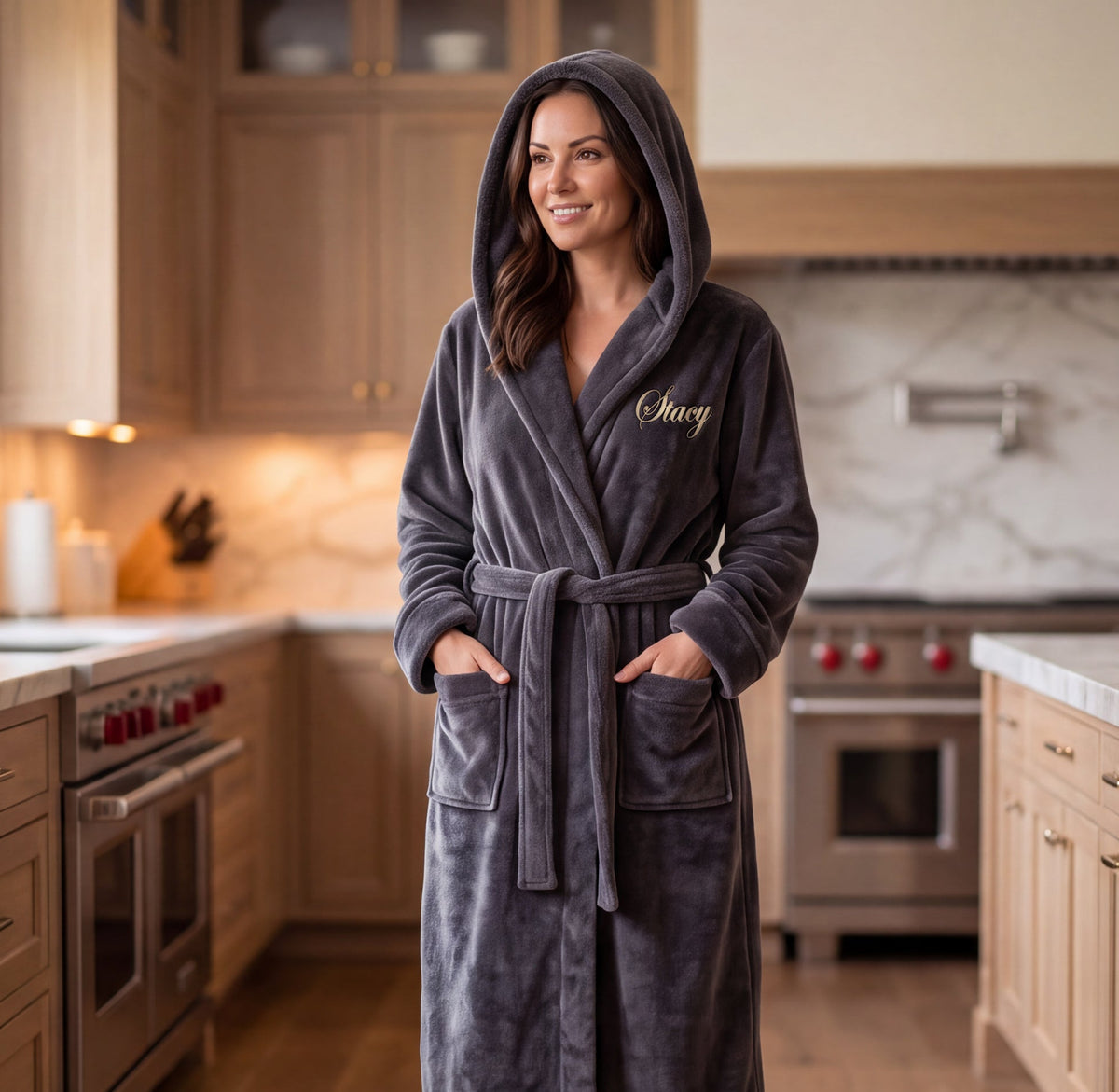 A woman stands comfortably in a modern kitchen wearing a dark gray plush hooded robe with a tie belt and embroidered gold script on the chest.