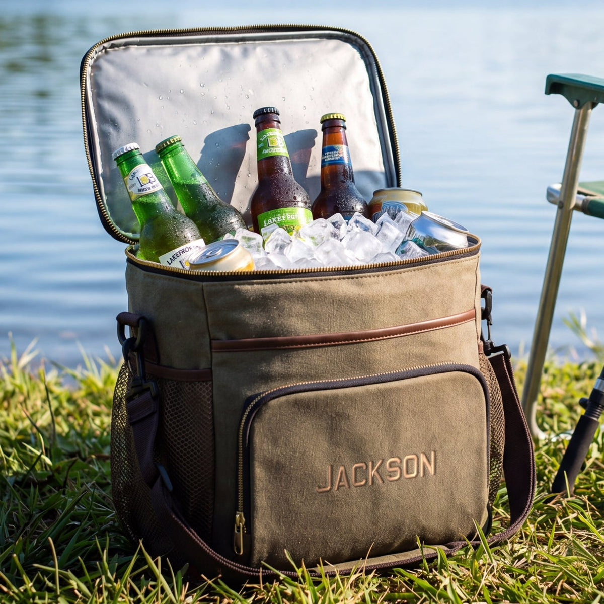 An open tan insulated cooler bag with the brand name JACKSON on the front pocket, filled with ice and beer bottles, sitting on grass by a lake. A green folding camp chair and fishing rod are beside it, with water and trees in the background.