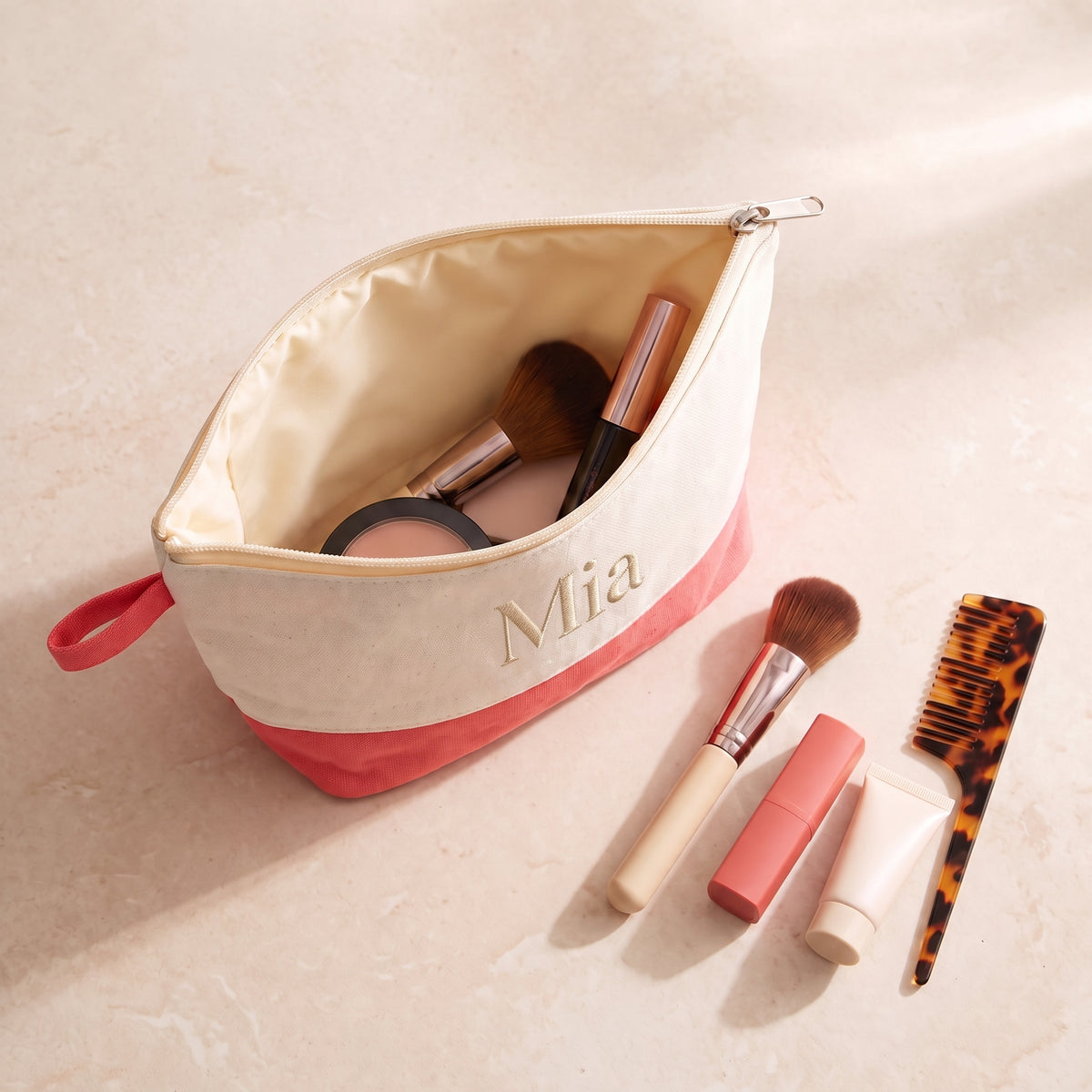 Overhead view of an open cream and coral-pink cosmetic bag embroidered Mia, showing its lined interior and makeup essentials.
