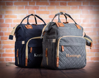The Nursery Run Diaper Backpack — quilted navy and gray, embroidered with the family names Keating and Hastings