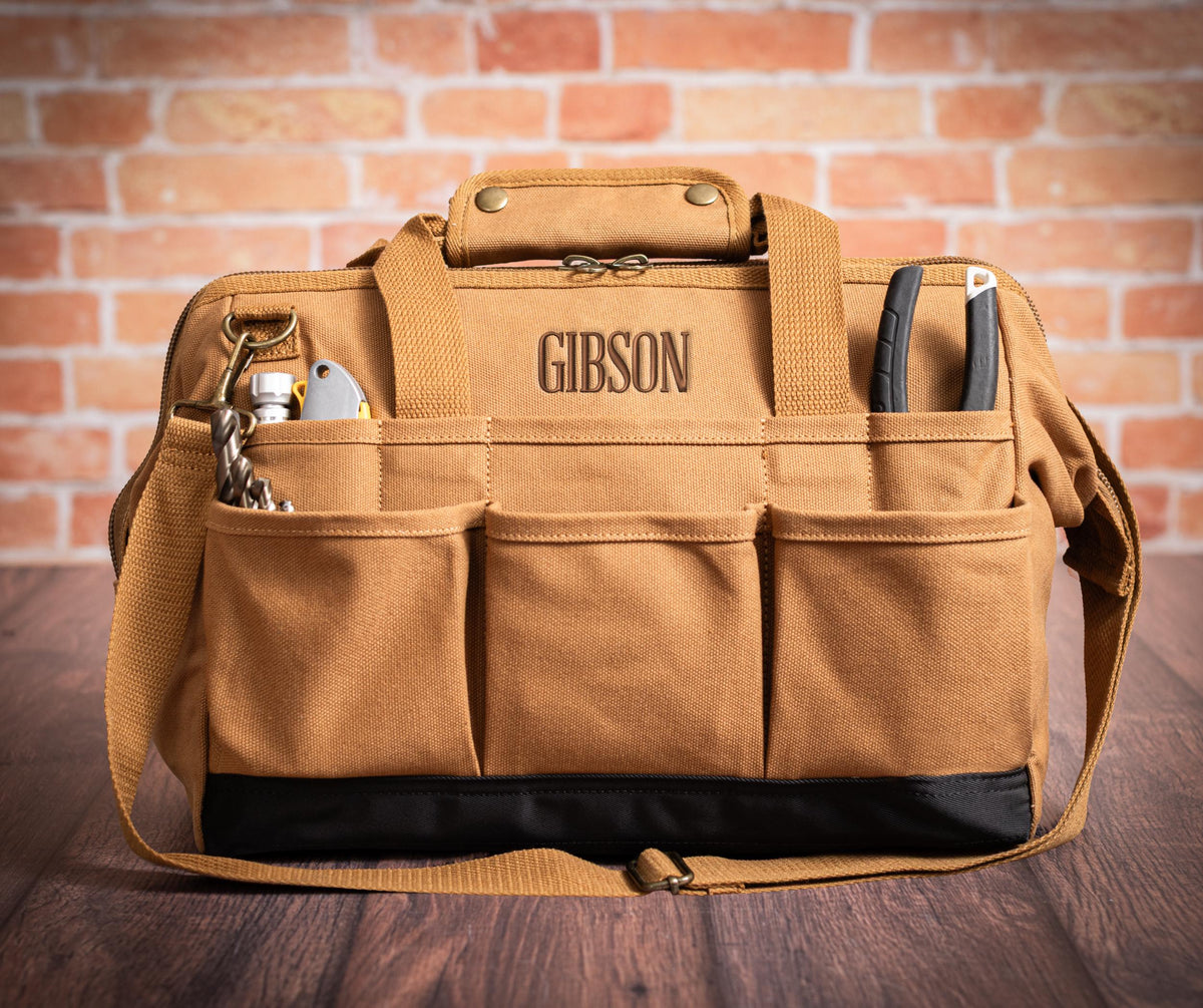 Brown canvas tool bag embroidered Gibson, with layered front pockets, shoulder strap and black base.