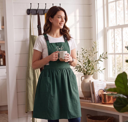 Juniper Coffeehouse pinafore apron with pink embroidered initials, worn in a bright kitchen while holding a mug.