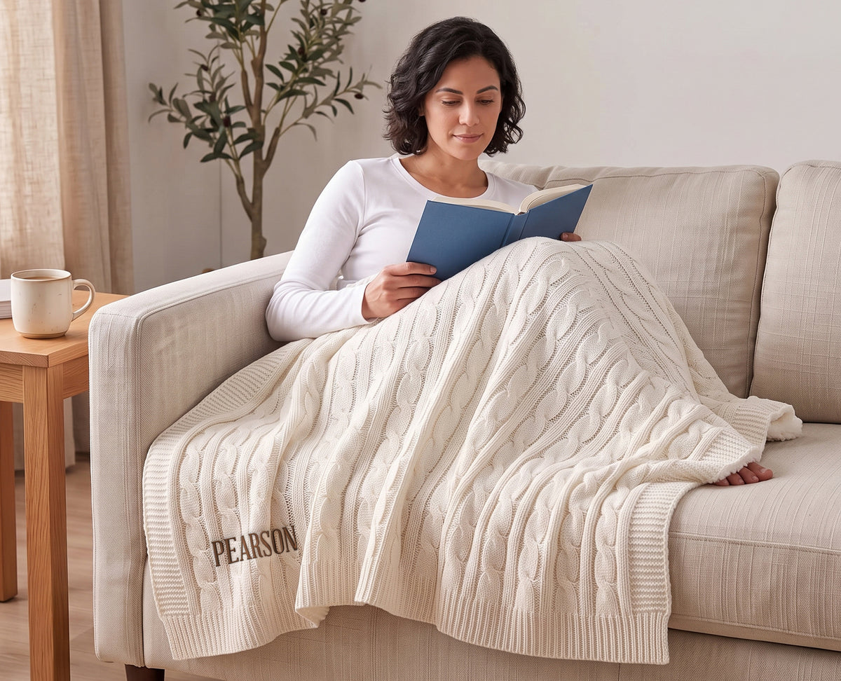 Woman reading on a sofa under a Cream cable-knit throw embroidered with the family name Pearson.