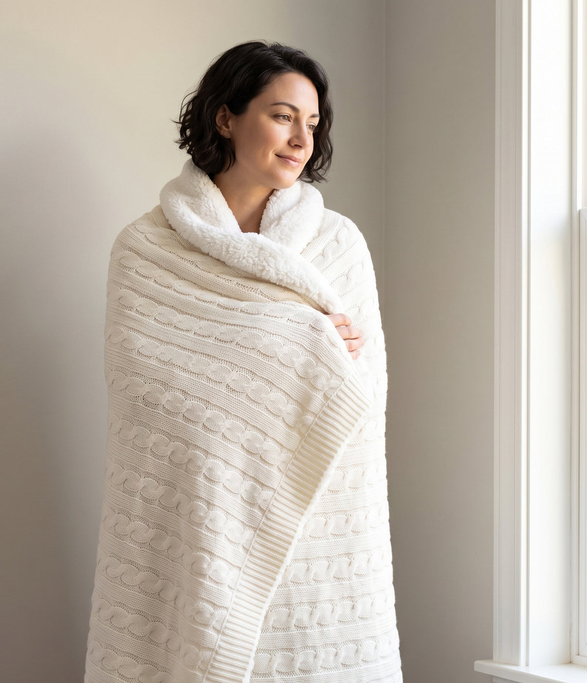 Woman wrapped in a Cream cable-knit throw with white sherpa backing beside a sunlit window.
